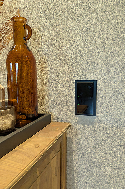 Een zwart custom-made wandpaneel van Grenton. Het verticale rechthoekige paneel bestaat uit 8 vlakken, opgedeeld in twee rijen van vier. In de buitenhoek van elk vierkant zie je een klein groen vierkant lichtje. Het paneel is bevestigd op een witte gespachtelde muur naast een eikenhouten dressoir. Op het dressoir staat een antraciet-grijze houten bak met daarin verschillende glazen vazen in bruin- en grijstinten.