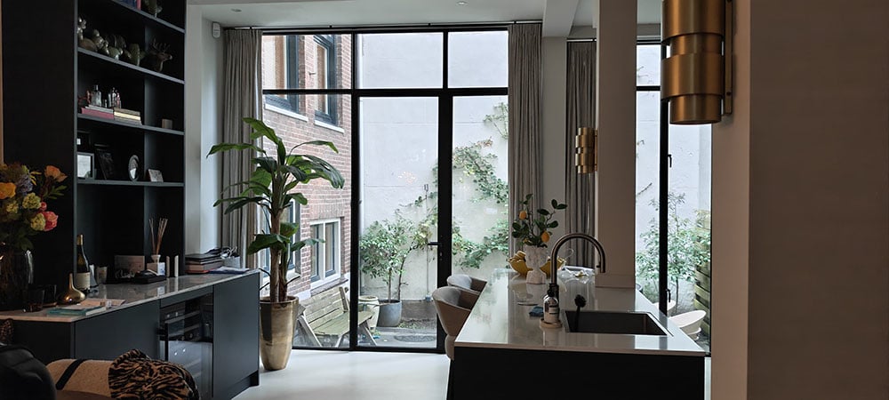 Moderne zwarte keuken in amsterdamse pijpenla. Kasten matzwart. Links een aanrecht met beige marmer en een wijnkoelkast. Plafondhoge ramen met uitkijk op kleine binnentuin. In linkerhoek een grote groene kamerplant. Boven het aanrecht een open zwarte kast met planken met daarop snuisterijen.