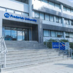 Grijs gebouw van de Frederick University op Cyprus. Voor de ingang met vier glazen deuren een brede trap bestaande uit 13 treden. Deel van het EU project INBLANC duurzaam wonen en werken waaraan Comfortica deelneemt.